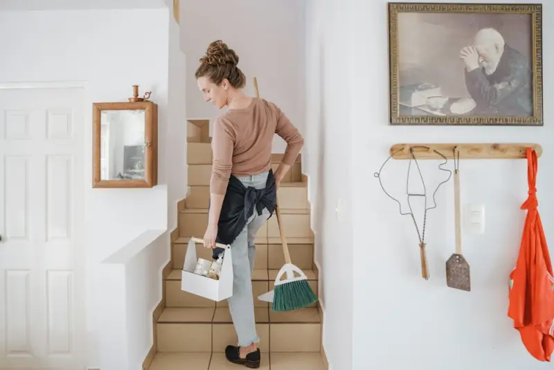 A cleaner carrying a caddy and a broom up the stairs of a home.