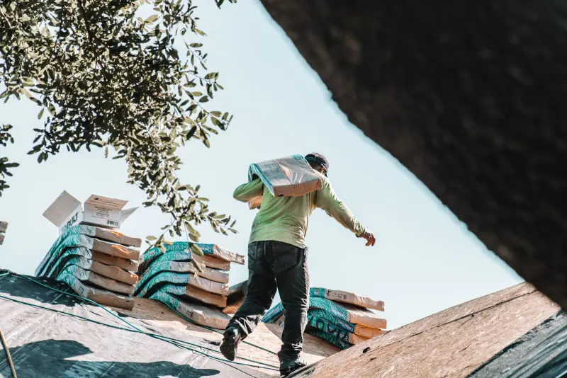 A roofer carrying a bundle of shingles across a roof.